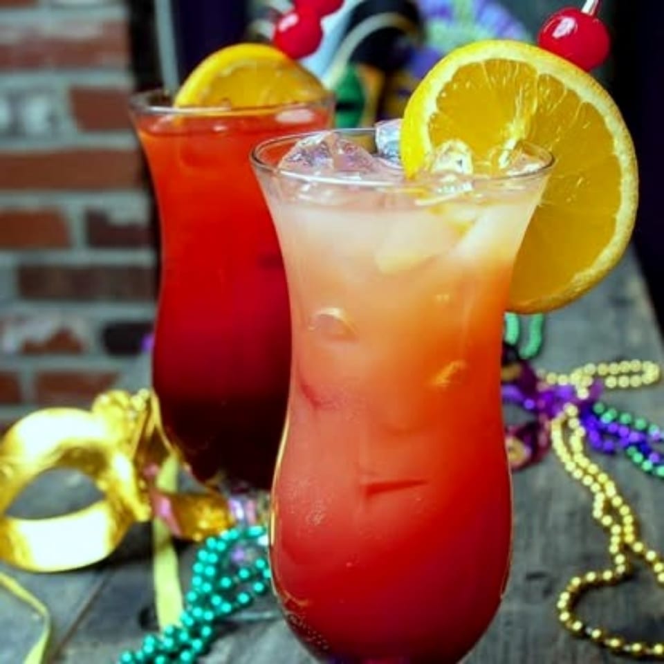 Two colorful fruit cocktails garnished with orange slices and cherries sit on a decorated table against a brick wall.