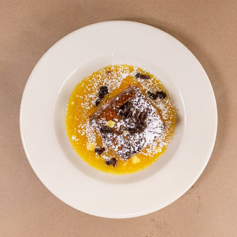 Bourbon St. Cafe's bread pudding with golden sauce, raisins, and powdered sugar on a white plate.