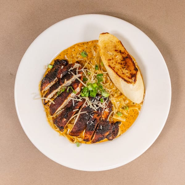Blackened chicken pasta with creamy sauce, green onions, shredded cheese, and toasted bread on a white plate.