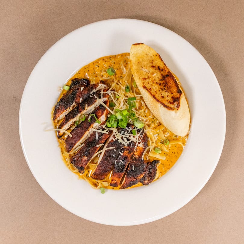 Blackened chicken pasta with creamy sauce, green onions, shredded cheese, and toasted bread on a white plate.