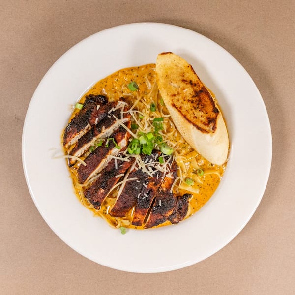 Blackened chicken pasta with creamy sauce, shredded cheese, green onions, and toasted bread on a white plate.