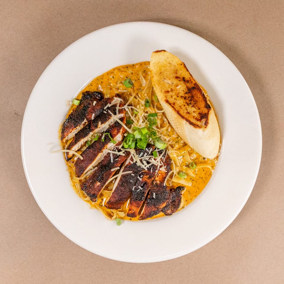 Blackened chicken pasta with creamy sauce, shredded cheese, green onions, and toasted bread on a white plate.