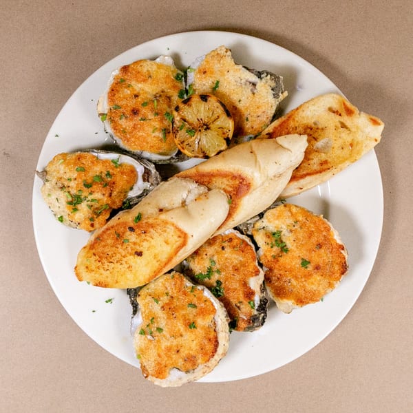 A white plate holds baked oysters, toasted bread, and a grilled lemon half, garnished with parsley.