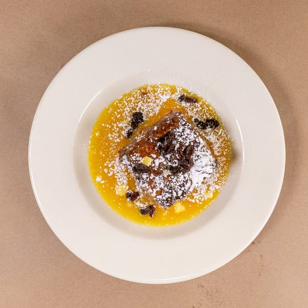 Bread pudding with yellow sauce, powdered sugar, and raisins on a white plate.