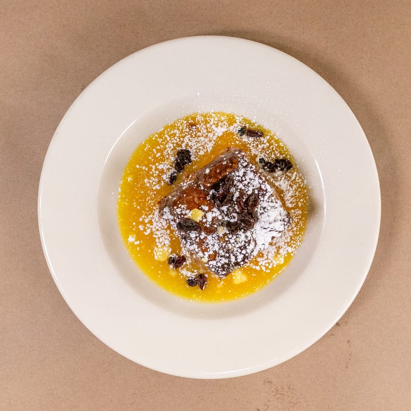 Bread pudding with yellow sauce, powdered sugar, and raisins on a white plate.