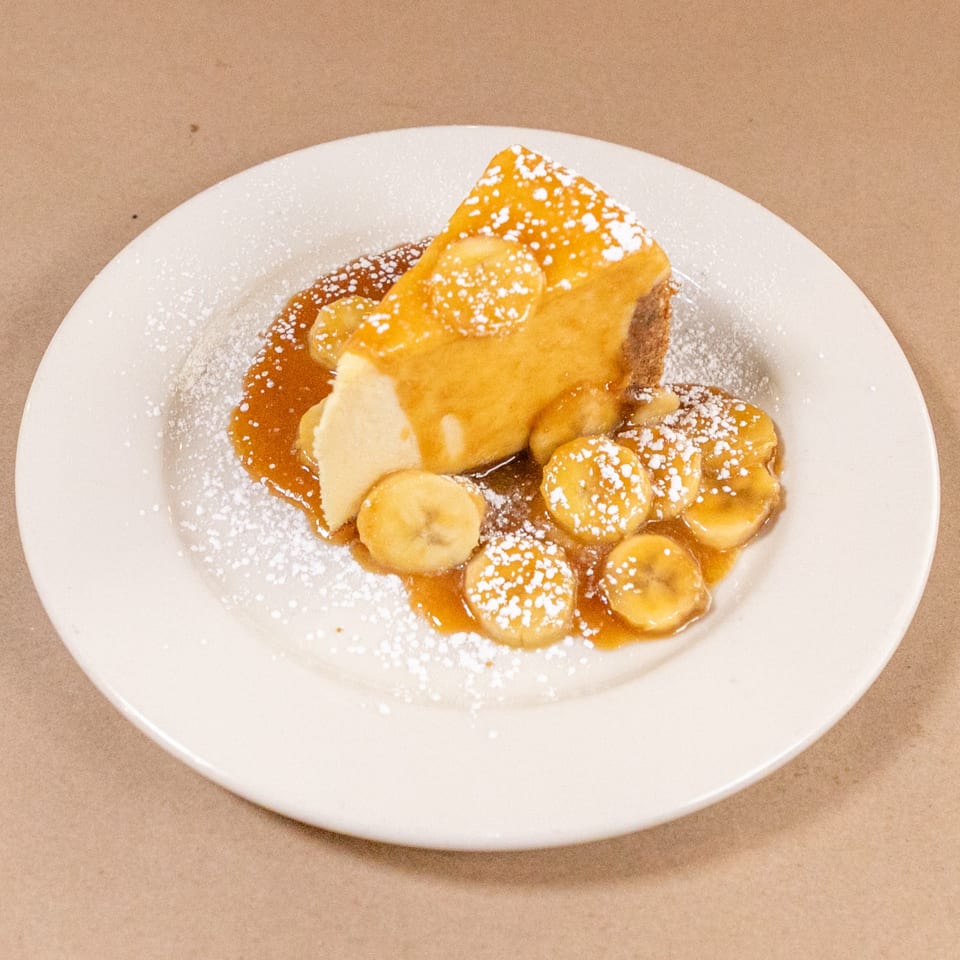 A slice of cheesecake topped with caramel, sliced bananas, and powdered sugar on a white plate.