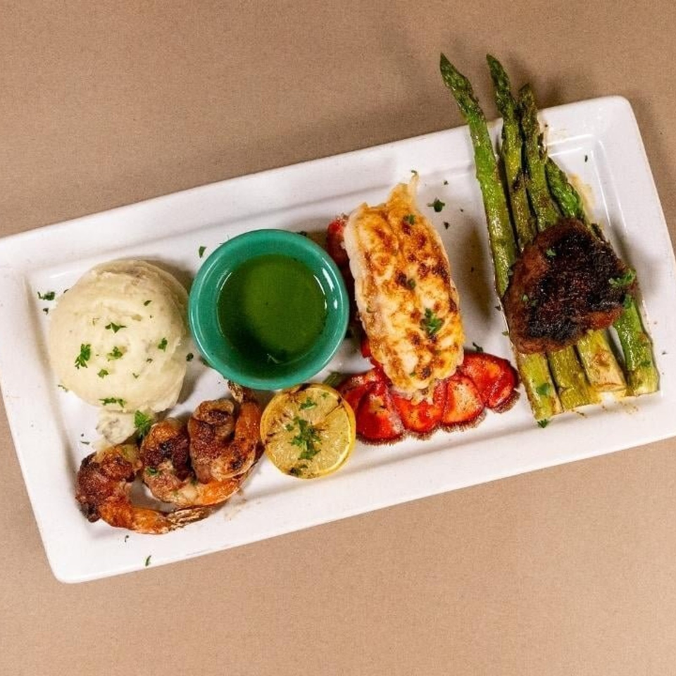 Grilled seafood platter with shrimp, lobster tail, scallops, asparagus, mashed potatoes, lemon, and green dipping sauce.