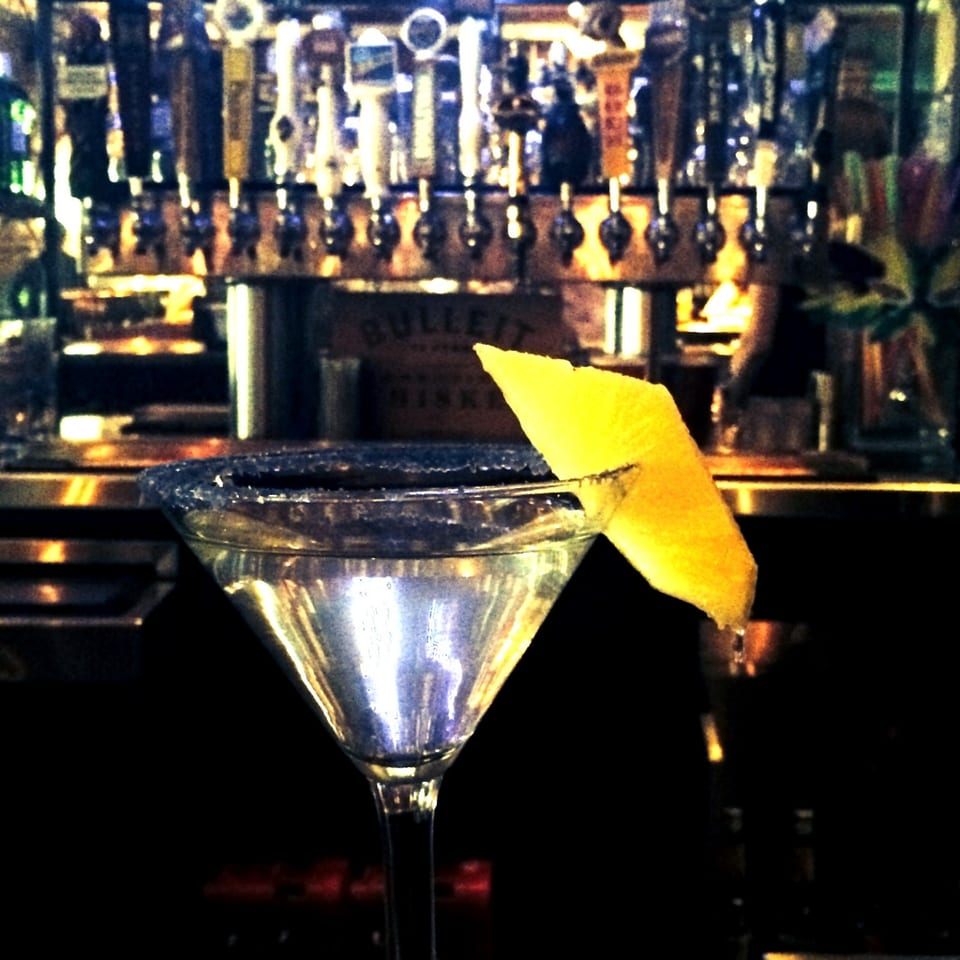 Martini glass garnished with a lemon twist sits on a dark bar, with liquor bottles and warm lighting in the background.