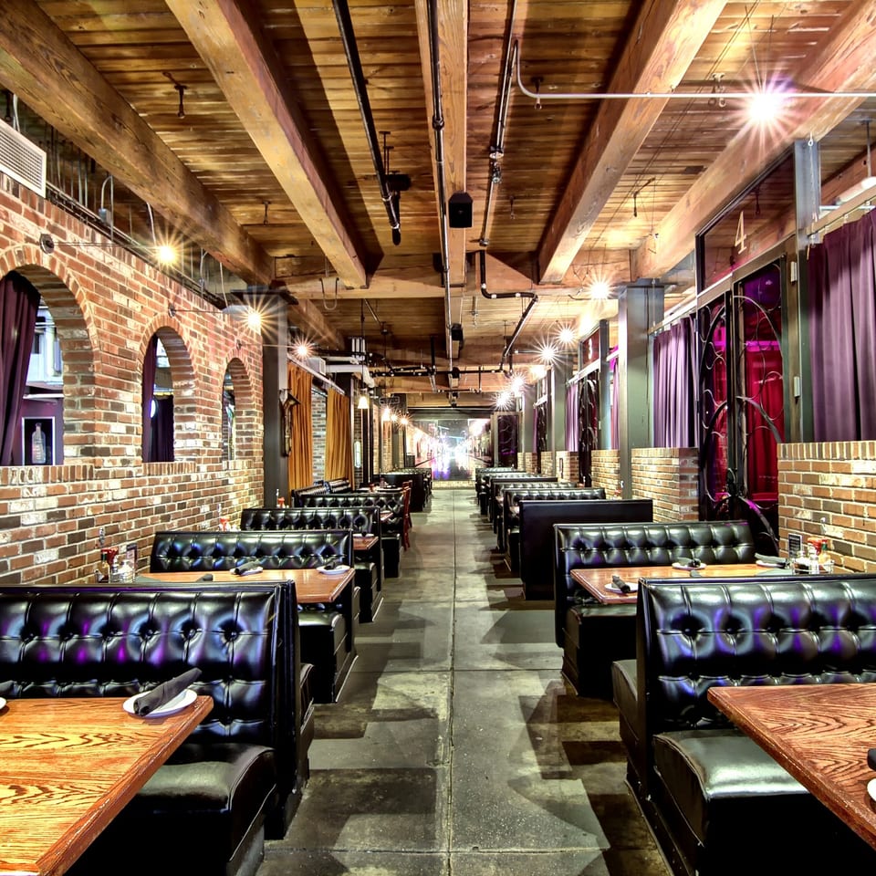Industrial-style restaurant dining room with brick walls, exposed wooden beams, black leather booths, and warmly lit tables.