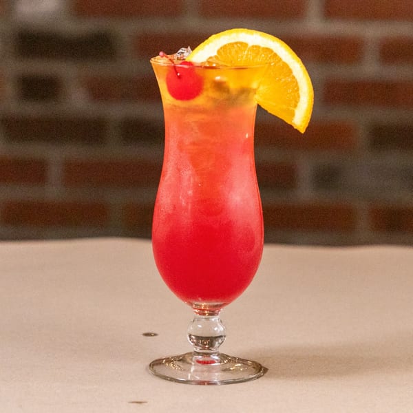 A vibrant red and orange cocktail, garnished with a cherry and orange slice, sits on a table.