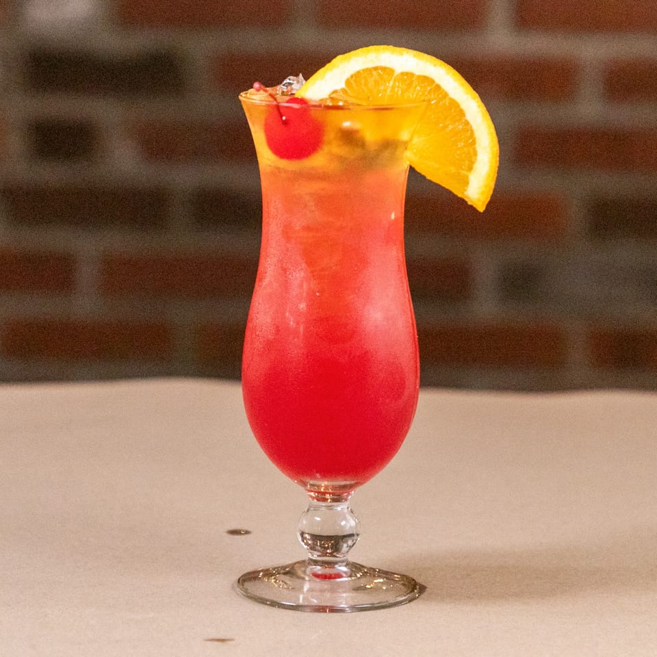 A vibrant red and orange cocktail, garnished with a cherry and orange slice, sits on a table.