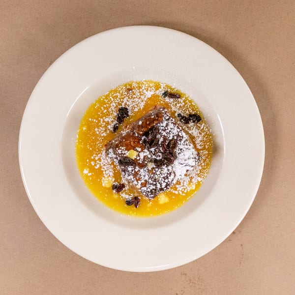 Bread pudding with orange sauce, powdered sugar, and raisins on a white plate.