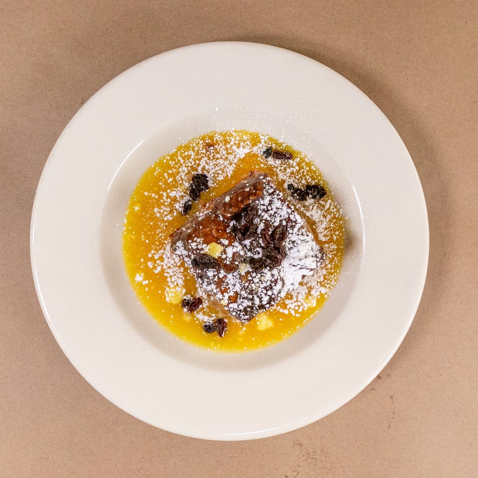 Bread pudding with orange sauce, powdered sugar, and raisins on a white plate.