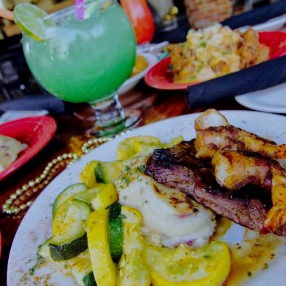 Colorful restaurant spread featuring grilled meat, roasted vegetables, pickles, sauces, and a bright green cocktail.