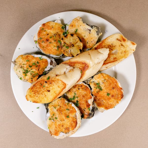 A white plate holds baked oysters topped with breadcrumbs and parsley, alongside grilled lemon and toasted bread.