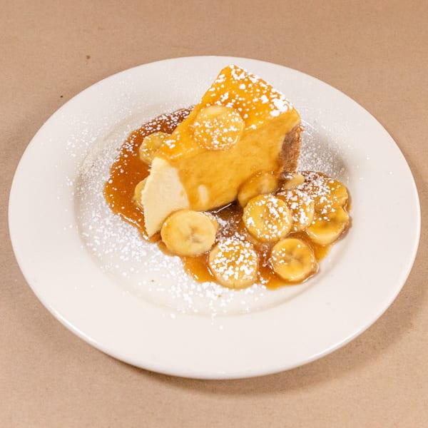 A slice of cheesecake topped with caramel, banana slices, and powdered sugar on a white plate.