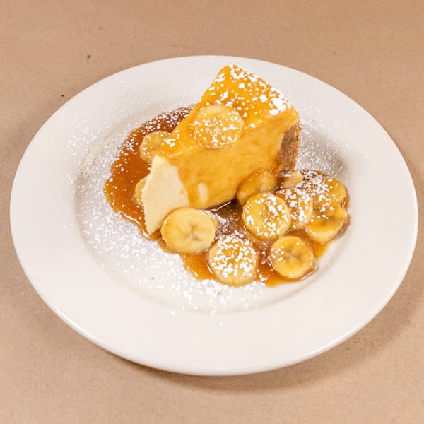 Slice of Bourbon St. Cafe cheesecake topped with caramel, banana slices, and powdered sugar.
