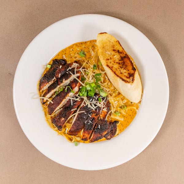Blackened chicken pasta with creamy sauce, green onions, shredded cheese, and toasted bread on a white plate.
